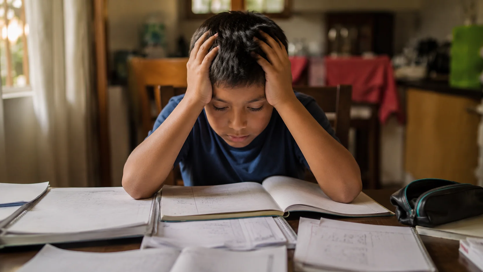 Niño ecuatoriano de unos 9 años sentado a la mesa de su comedor rodeado de libros y cuadernos abiertos, apoyando la cabeza en sus manos con expresión de cansancio y agobio bajo una luz cálida de la tarde